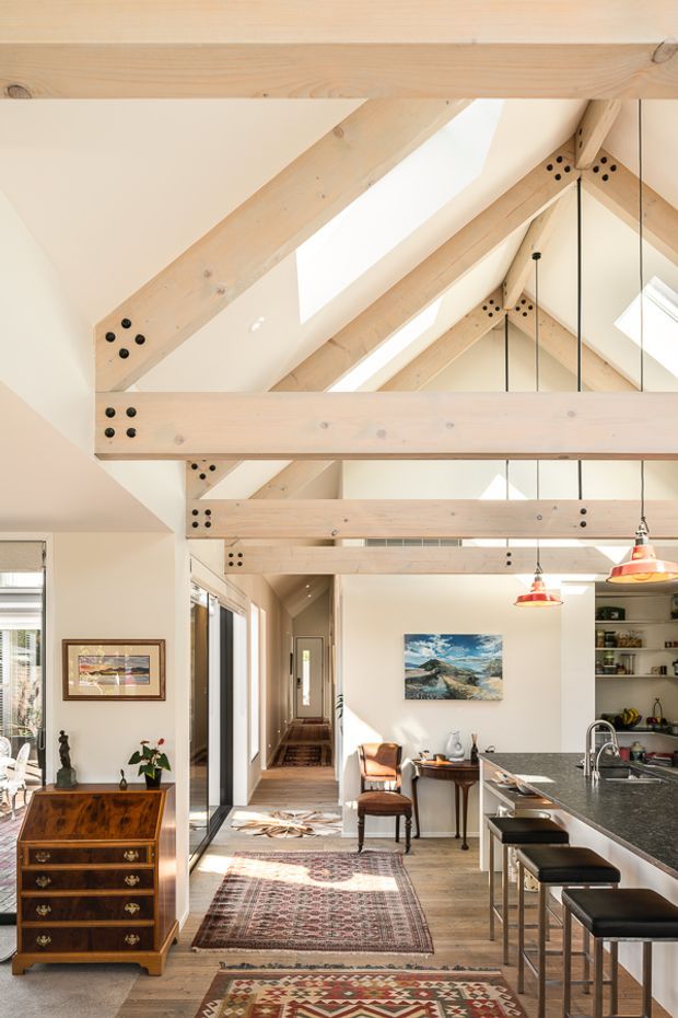 Bright open-plan interior featuring a kitchen and dining/living area beneath a vaulted ceiling with exposed pale timber beams and visible structural connections. The space includes a long dark kitchen island with bar seating, pendant lights, timber flooring, patterned rugs, traditional furniture, and skylights that bring in abundant natural light. A hallway and glazed openings extend toward the rear.