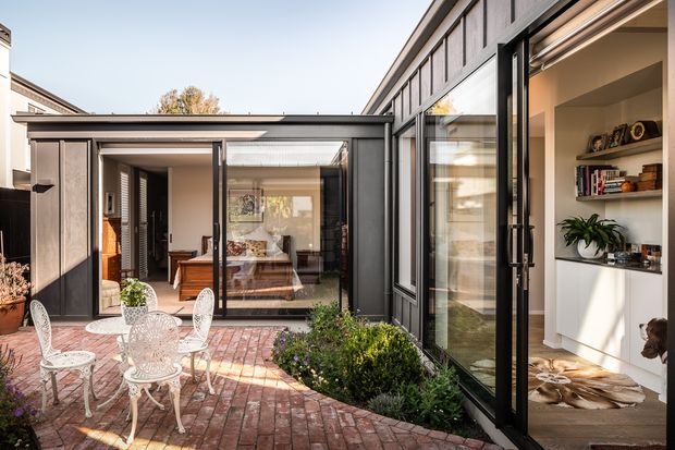 A bright courtyard view of a contemporary home extension, featuring a red brick-paved outdoor area with white metal café furniture, dark framed floor-to-ceiling glazing, and a connected indoor living space with built-in shelving. The design opens the interior directly onto the courtyard and includes a low flat-roofed glazed wing.