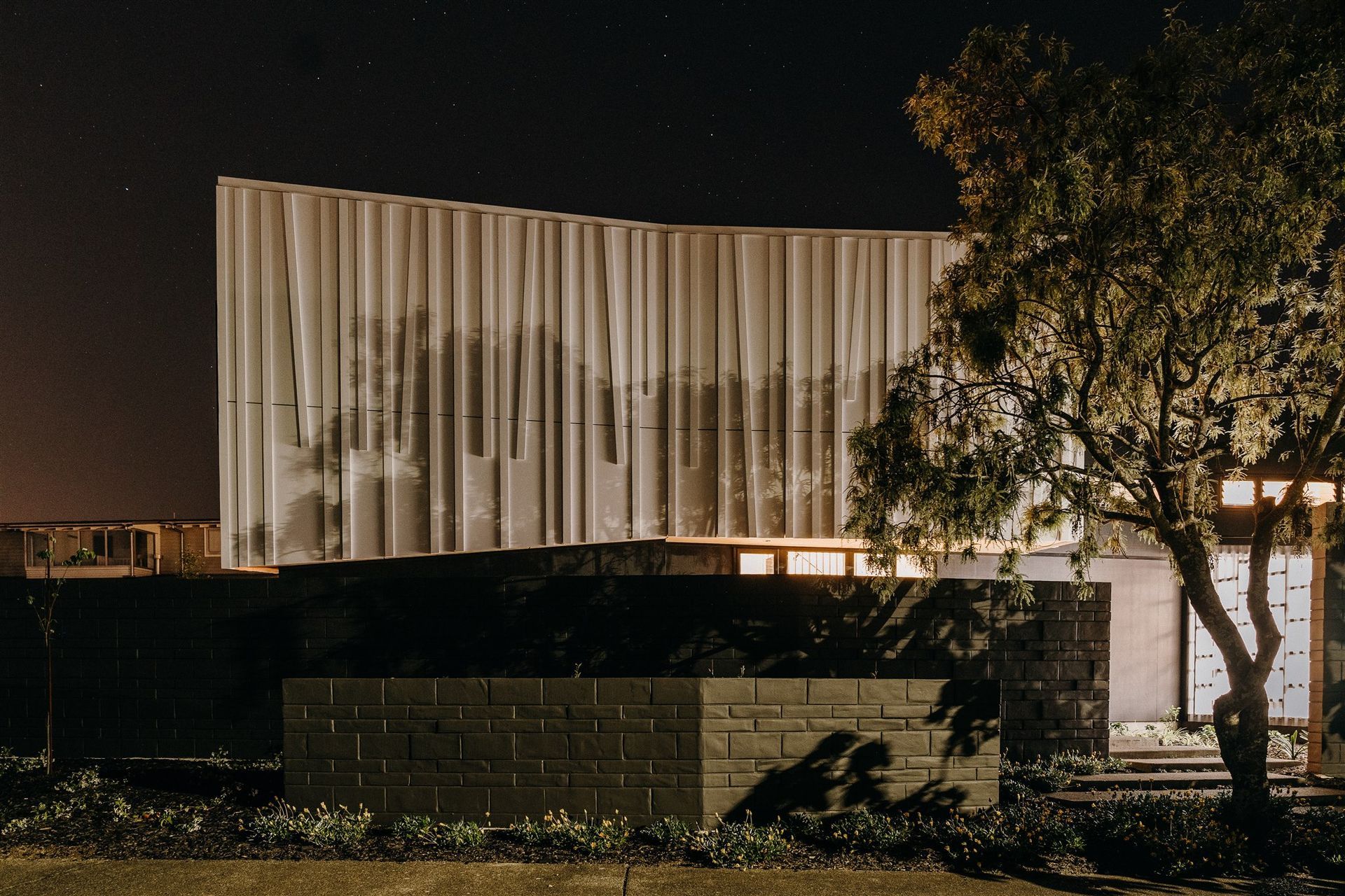 The 'shroud' or top level of the house is illuminated at night, with shadows cast from the surrounding trees to highlight the unique textural form.