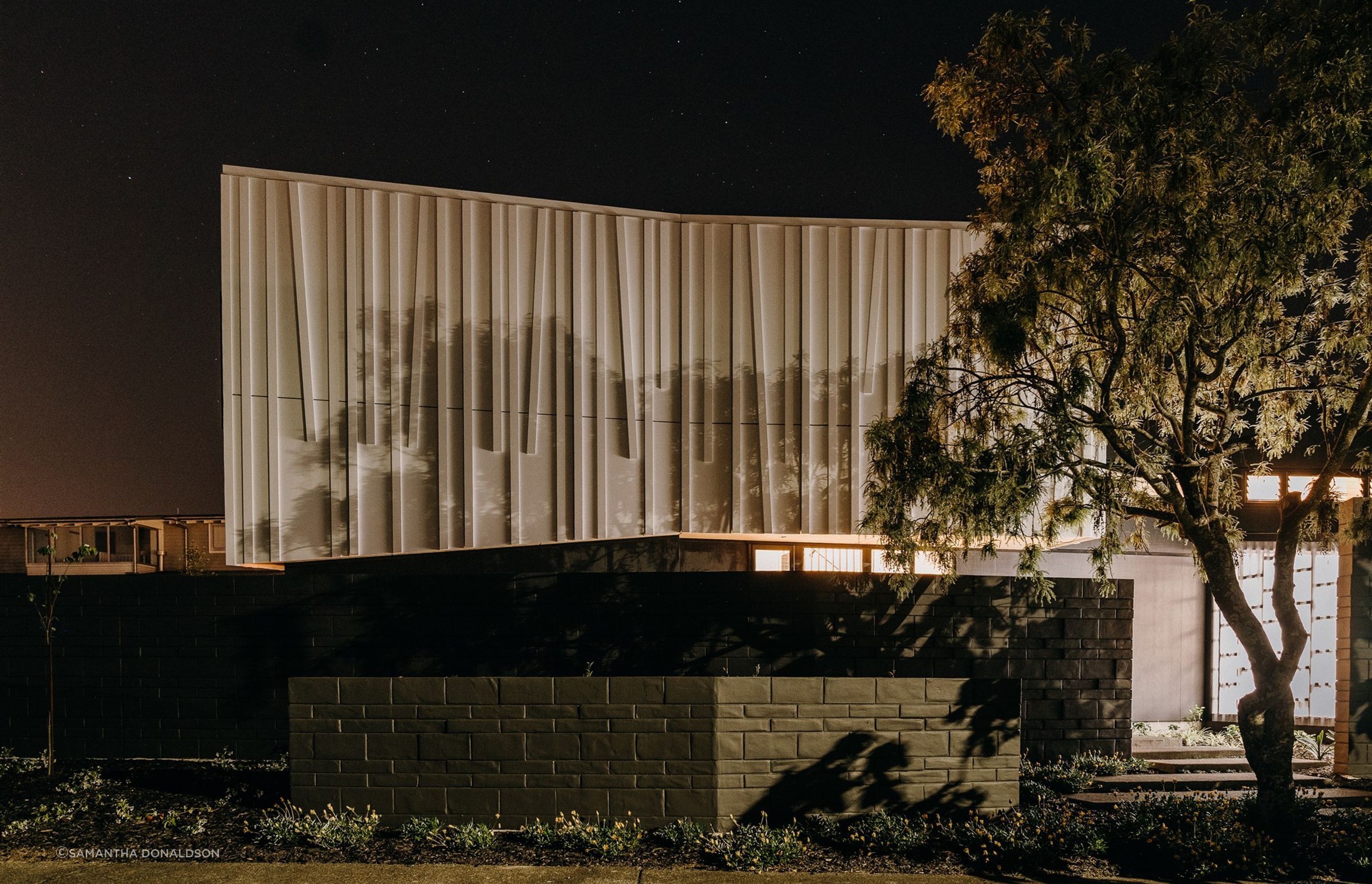 The 'shroud' or top level of the house is illuminated at night, with shadows cast from the surrounding trees to highlight the unique textural form.
