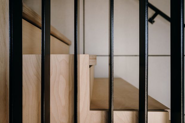 Close-up architectural detail of a timber staircase or stepped built-in joinery viewed through a series of slim black vertical metal balusters. Pale natural timber treads and cabinetry-like panels contrast with a light neutral wall, while a black ceiling-mounted light fitting is partially visible in the background. The image is tightly framed around the stair and balustrade, with a refined contemporary material palette.