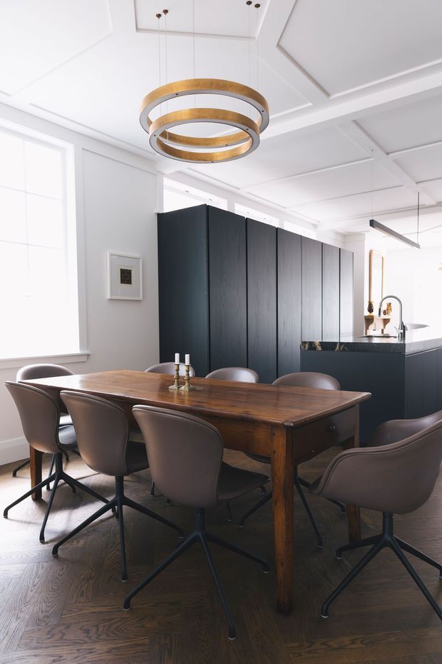 A bright, refined open-plan dining and kitchen interior. A substantial dark timber dining table is surrounded by sculptural brown upholstered chairs, positioned on dark timber flooring. Behind it is a dramatic matte-black kitchen with a large island, while white walls, tall windows, and a detailed coffered ceiling create a light contrast. A layered circular pendant in timber and metallic tones hangs above the dining table.