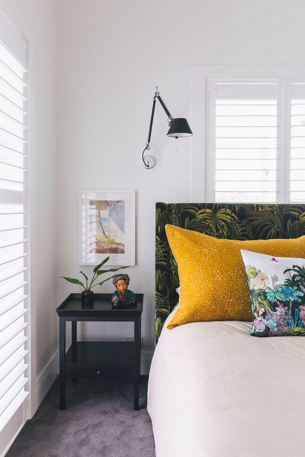 A bright, compact bedroom corner with white walls, plantation shutters, grey carpet, and a bed featuring a bold dark botanical upholstered headboard. Mustard and floral patterned cushions add colour. A black bedside table holds a small plant and decorative object, while a black wall-mounted adjustable lamp and framed artwork complete the room.