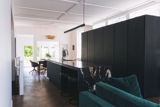 A bright open-plan kitchen and living area in a refined contemporary interior. The kitchen features a large dark island with a dramatic veined stone waterfall edge, matching dark floor-to-ceiling cabinetry, black pendant lighting, and bar stools. A teal sofa occupies the foreground, while a dining area and garden views are visible beyond. The room has white coffered ceilings, tall windows, and dark timber or wood-look flooring.