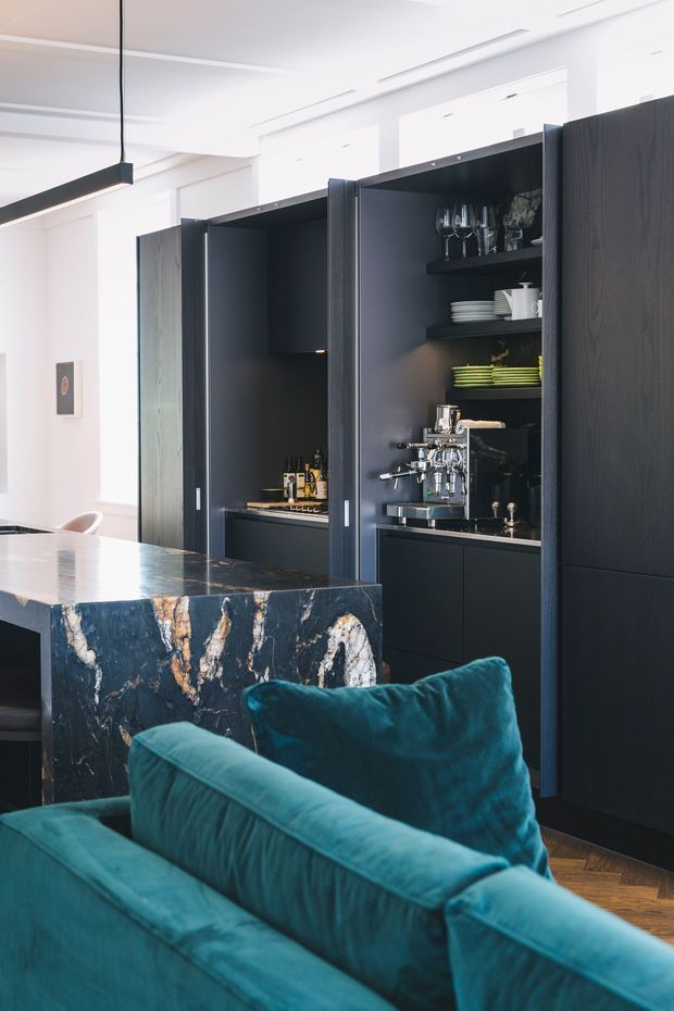 A sophisticated open-plan interior featuring a dark timber drinks and coffee-bar cabinetry wall with open shelving, glassware, crockery, bottles and an espresso machine. A dramatic dark veined stone island or peninsula forms the foreground, with a deep teal upholstered sofa and herringbone timber flooring. The bright white ceiling and walls contrast with the dark joinery and rich blue-green furnishings.