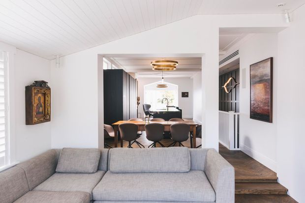 Bright open-plan interior showing a living room in the foreground with a large light-grey sectional sofa, opening onto a dining area with a timber table and upholstered chairs. White-painted walls and timber-lined ceiling create an airy backdrop, while dark built-in cabinetry, timber flooring, contemporary pendant lighting and decorative wall art add contrast. A small change in floor level and a partially visible stair or raised passage appear to the right.
