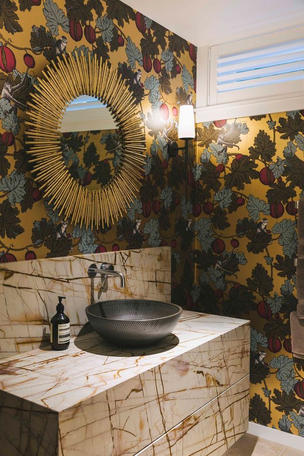 A richly decorated small bathroom featuring dramatic botanical wallpaper with dark foliage, fruit, and bird motifs. A large ornate gold sunburst mirror hangs above a marble-clad vanity with an integrated raised basin, wall-mounted chrome tapware, and a contemporary wall sconce. A high horizontal window with white louvred blinds provides natural light. The scheme is opulent, theatrical, and highly patterned.