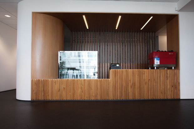 A minimalist reception or service counter set within a large recessed timber-lined opening. Vertical timber slats form the front counter and rear feature wall, with warm wood side panels and slim linear recessed ceiling lights. A glass display unit and red equipment sit on the counter. The surrounding architecture is crisp white with a dark floor, creating a clean, contemporary commercial interior.