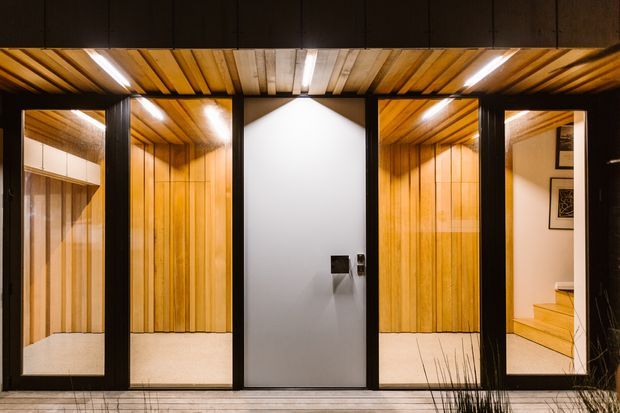 A dark-framed contemporary interior with a series of timber-lined recessed niches or rooms opening onto a tiled foreground. Warm vertical timber wall and ceiling panelling contrasts with a central smooth white wall containing two dark fixtures. The right-hand opening reveals a small set of timber stairs and framed artwork, while the left side includes pale built-in cabinetry and a reflective surface. Linear integrated lighting emphasizes the timber ceiling.