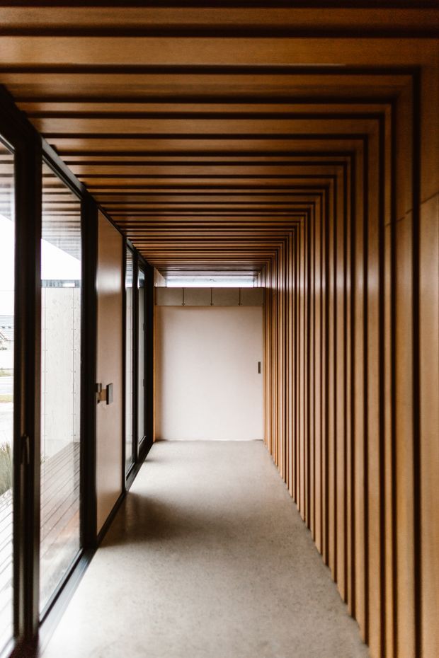 A long, narrow architectural corridor with full-height dark-framed glazing on the left and a continuous warm timber-lined wall, ceiling, and structural rib detail on the right and overhead. The repeated timber battens create a strong perspective tunnel effect, while the pale polished concrete or terrazzo-like floor and white end wall provide contrast. The space is empty, contemporary, and naturally lit from the glazing.