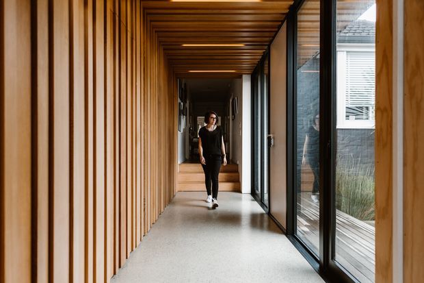 A long, refined interior hallway or gallery corridor with warm vertical timber wall battens on the left and a matching timber slatted ceiling. Full-height black-framed glazing runs along the right, opening views to a landscaped courtyard and an adjacent dark-clad building. The corridor has a light polished floor, recessed linear lighting, and a short set of steps leading toward a darker interior beyond. A person provides scale.