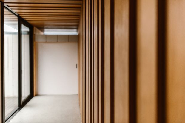 A narrow, minimalist interior passage or landing with full-height black-framed glazing on the left, a warm timber slatted ceiling overhead, and a prominent vertical timber batten wall on the right. Pale flooring and white walls create a calm, refined corridor-like space.