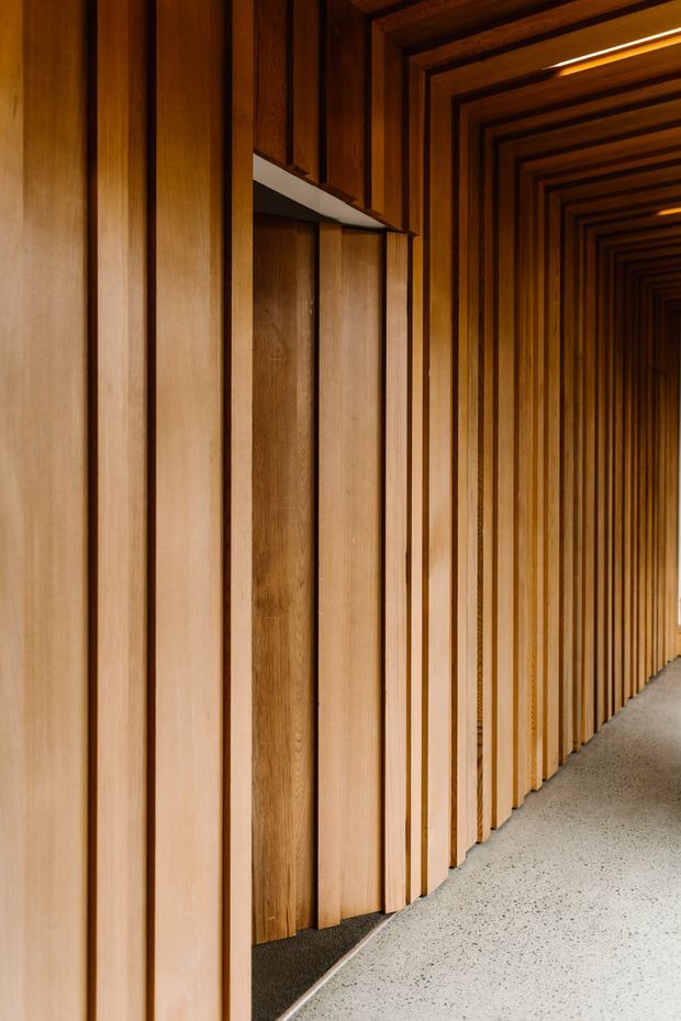 A long, narrow interior corridor lined with rhythmic vertical timber wall battens and deep timber door or wall recesses. Warm concealed linear lighting highlights the layered wood detailing, while a speckled light terrazzo or concrete floor curves gently along the passage. The image focuses on the refined timber-lined architectural treatment and strong perspective.