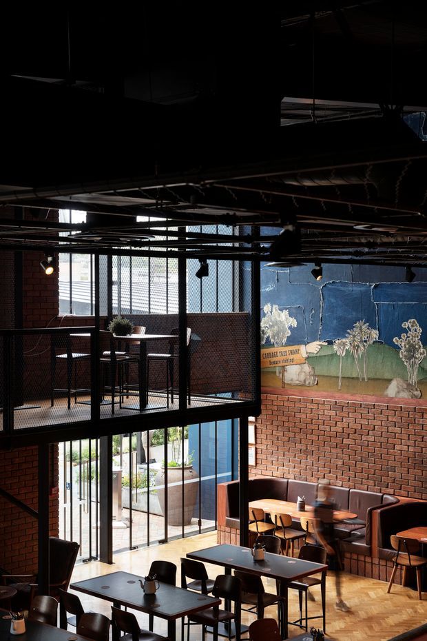 A dark, industrial-style hospitality interior, likely a café or restaurant, with exposed ceiling services and structure, red brick walls, black metal mesh screens and framing, timber parquet flooring, booth seating, dining tables, and a large painted mural. Daylight enters through tall glazed openings, with a mezzanine level overlooking the dining area.