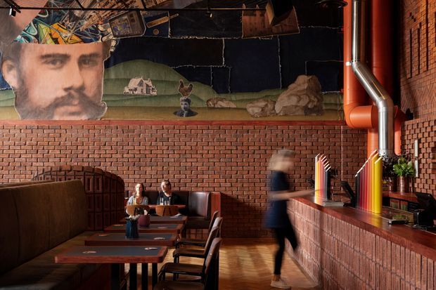 A moody, eclectic commercial interior resembling a bar or restaurant, with exposed red brick walls, warm timber flooring, a long brick-faced counter, industrial metal ductwork, dark booth seating, and timber tables and chairs. A large surreal mural and suspended artwork decorate the upper wall and ceiling, while blurred patrons and staff create a lively atmosphere.