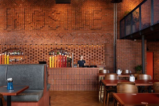 A warm, industrial-style commercial dining and bar interior with exposed red brick walls, decorative brickwork, a tiled or brick bar front, timber herringbone flooring, dark metal mezzanine railings, and rows of wooden tables and chairs. A staffed bar with colourful beverage dispensers and bottles occupies the rear, while booth seating and dining tables fill the foreground.