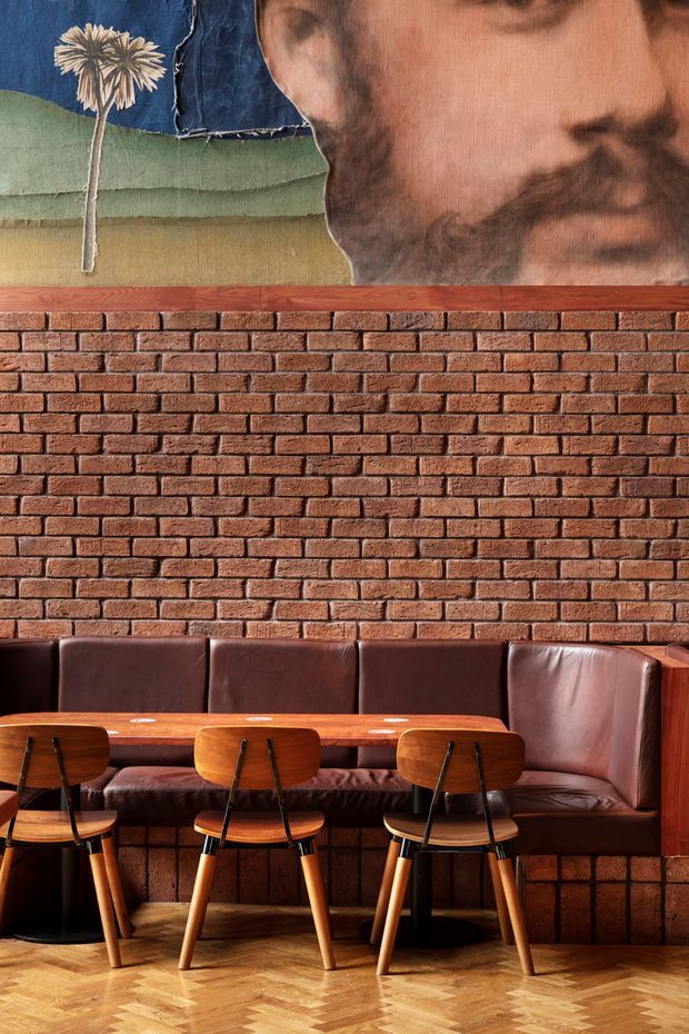 A stylish restaurant or café dining area with a long built-in brown leather banquette, a polished timber dining table, and several mid-century-inspired wooden chairs. The space features an exposed red brick wall, warm herringbone timber flooring, dark timber joinery, and a large vintage-style artwork above the banquette. The overall atmosphere is warm, richly textured, and retro-industrial.