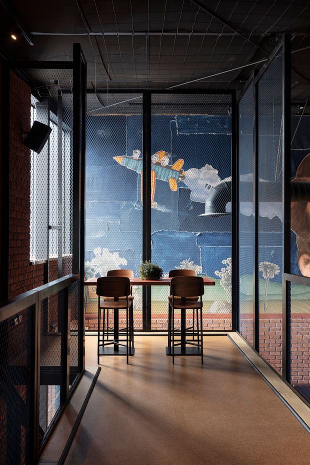A dark, industrial-style interior with a narrow mezzanine or enclosed walkway overlooking a vibrant muralled space. Black metal mesh partitions and framing surround a small bar-height timber counter with two dark timber stools. Exposed brick walls, dark gridded ceiling, metal details, and warm flooring create a moody hospitality or commercial atmosphere.