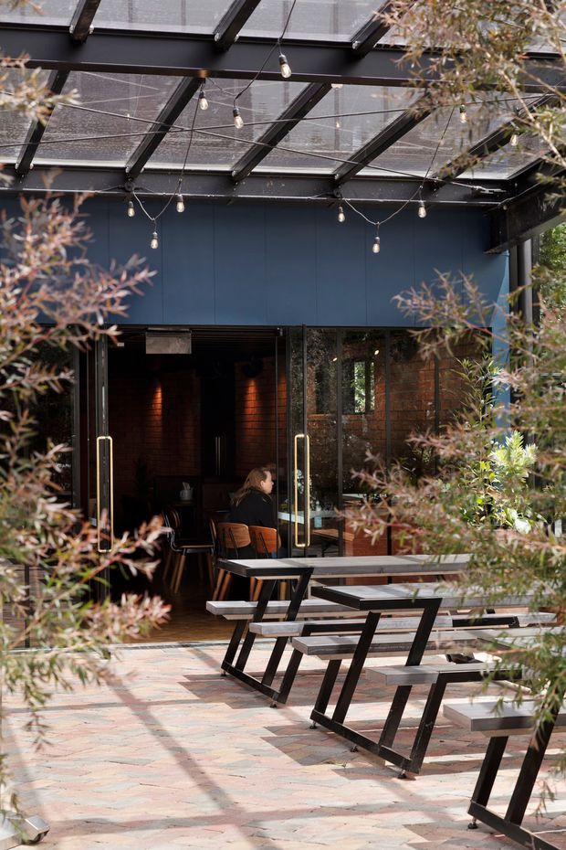 A covered outdoor dining terrace in front of a dark, modern venue. The space features a transparent roof canopy supported by black metal framing, festoon lights, large glazed folding doors, a dark blue facade, exposed brick visible inside, and long timber-and-black-metal picnic tables on a multicoloured brick-paved surface. Planting partially frames the foreground.