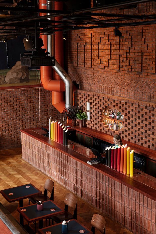 A moody commercial bar and dining interior with exposed red brick walls, a long brick-fronted bar counter, dark timber surfaces, colourful cylindrical bar accents, black metal services and ductwork, and herringbone timber flooring. Small tables and bentwood chairs occupy the foreground, while bottles and glassware are displayed behind the bar. The industrial structure is softened by warm lighting and rich red-brown materials.