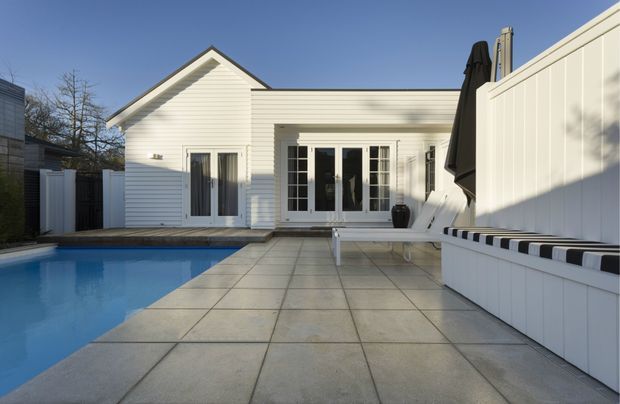 A bright white weatherboard-style single-storey building opens onto a paved pool terrace. The scene features a blue swimming pool, large pale stone or concrete pavers, white French doors and windows, a tall white privacy fence, and built-in white poolside bench seating with black-and-white cushions. The gabled roof form and clean white palette create a crisp contemporary coastal atmosphere.