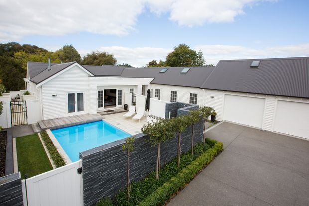 A single-storey residential property viewed from above, featuring white horizontal weatherboard cladding, a dark pitched metal roof with skylights, multiple garage doors, and a landscaped courtyard with a bright blue swimming pool, timber pool deck, and dark stone privacy wall.
