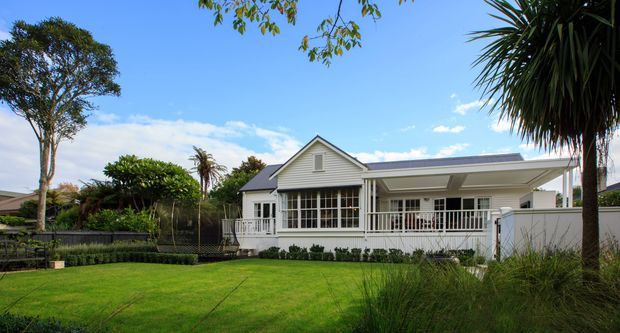 A single-storey suburban house viewed from the landscaped front yard. The home has a light weatherboard facade, a prominent gabled central section, a broad low-pitched roof extension, white-framed windows and doors, and a covered veranda with white balustrading. The property is surrounded by a manicured lawn, mature trees, ornamental grasses, and garden planting under a bright blue sky.