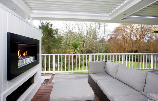 Covered outdoor lounge area with a white-lined ceiling, white vertical-panel feature wall, built-in fireplace, timber decking, white balustrade, and a large grey outdoor sectional sofa with ottoman. The space opens onto a leafy garden setting.