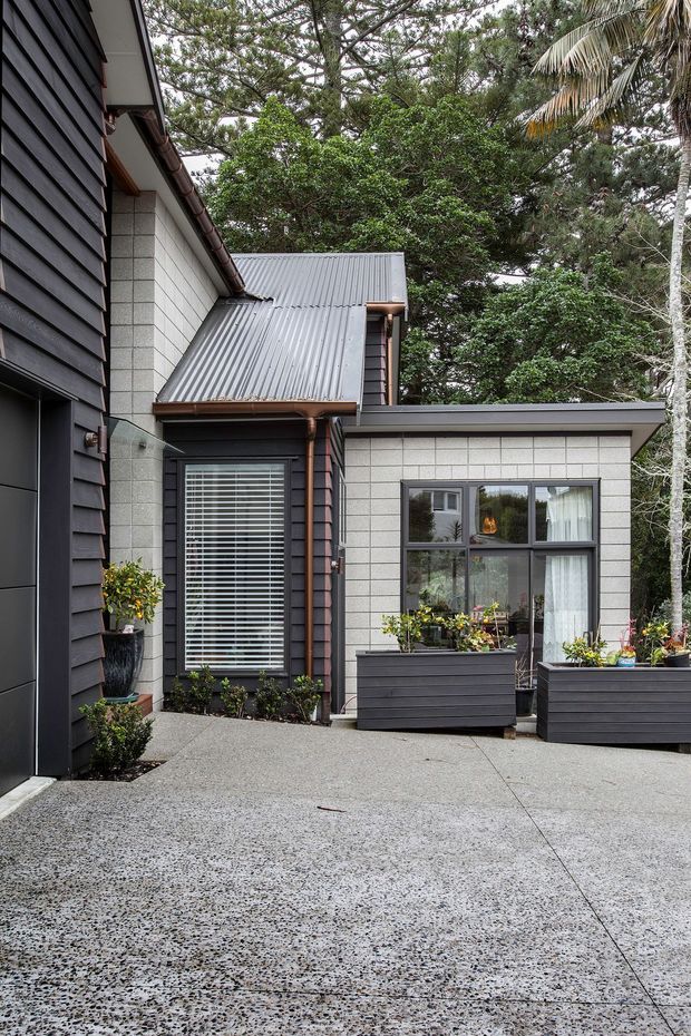 A compact courtyard-facing exterior of a contemporary house, featuring dark horizontal cladding, light grey block masonry walls, charcoal corrugated metal roofs, dark-framed windows, copper-toned gutters and downpipes, and raised dark planter boxes. The foreground is a broad textured concrete-paved courtyard, surrounded by mature trees and palms.