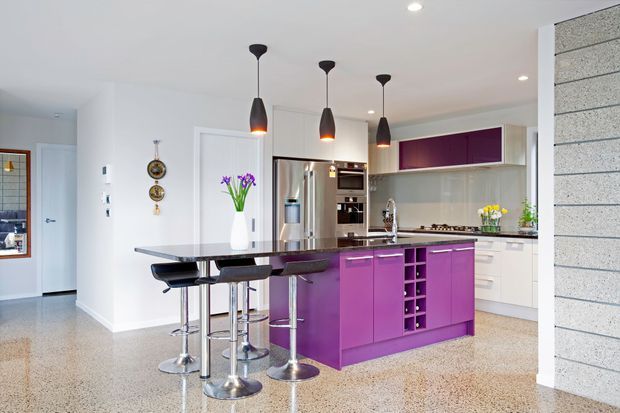 Bright contemporary kitchen with a vivid purple island and integrated wine storage, white cabinetry, stainless-steel appliances, a dark stone-look benchtop, and a matching breakfast bar with stools. Pendant lights hang over the island, while terrazzo-style flooring and a pale textured feature wall add material contrast.