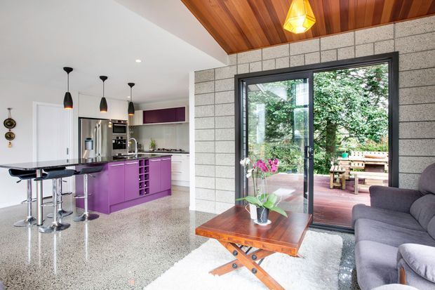 Bright open-plan interior combining a contemporary kitchen and living area. The kitchen features vivid purple cabinetry, stainless-steel appliances, a dark island benchtop with bar seating, and polished terrazzo-style flooring. The living zone has a grey sofa, timber coffee table, and a large black-framed glazed door opening to a planted outdoor area. A warm timber-lined sloped ceiling and exposed grey masonry feature wall add strong architectural character.