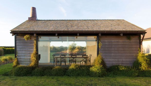 A compact single-storey detached garden building with weathered horizontal timber cladding, a low pitched tiled roof, and a broad glazed wall opening onto a dining area. A dark chimney rises from the roof, while climbing greenery and landscaping frame the structure. The rural setting and simple timber construction give it a cabin or outbuilding character.