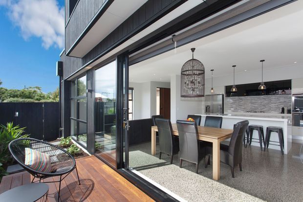 A bright contemporary open-plan dining and kitchen space opening onto a timber deck through large black-framed sliding glass doors. The room features a timber dining table with upholstered chairs, a white kitchen island with black stools, a textured grey splashback, pendant lighting, and a decorative birdcage pendant. Dark vertical exterior cladding and lush planting are visible outside.