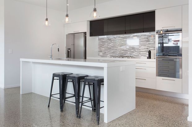 A bright contemporary kitchen with a large white island and breakfast overhang, three dark metal bar stools, integrated stainless-steel appliances, white cabinetry, dark upper cabinets, and a horizontal mosaic tile splashback. Polished aggregate or terrazzo-like flooring and minimalist pendant lights complete the space.