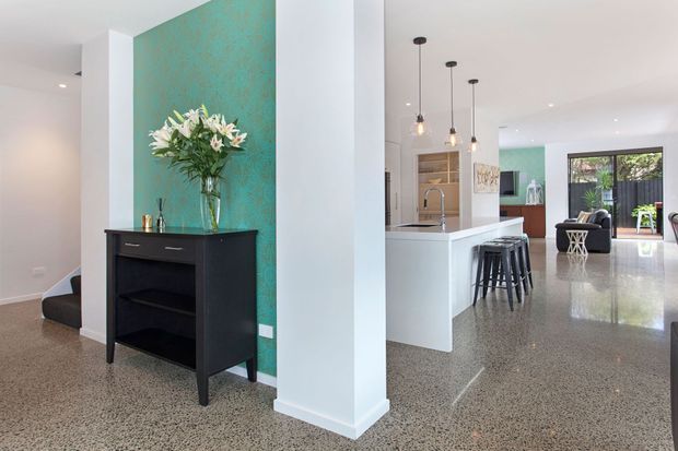 Open-plan contemporary home interior featuring a polished terrazzo floor, white walls and structural columns, a white kitchen island with black bar stools, and a living area beyond. A vivid turquoise feature wall with a black console and floral arrangement provides a strong accent. Pendant lights hang above the kitchen, while large glazed doors or windows connect the living space to greenery outside.