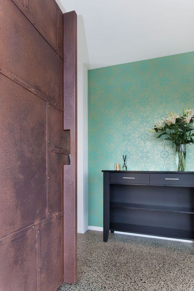 Interior entryway featuring a large rust-toned metal panel door with exposed fasteners, a vivid turquoise patterned feature wall, black built-in console cabinetry, and a polished terrazzo-style floor. A tall vase of flowers and small decorative objects sit on the console.