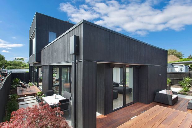 Modern dark-clad two-storey house with a sharp angular roofline, extensive glazing, and spacious timber decking around the exterior. Outdoor dining and lounge furniture occupy the deck, with landscaped planting visible around the edges.