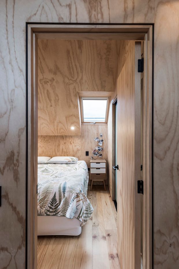 A compact bedroom viewed through an open timber doorway. The room is lined extensively in warm natural plywood, with matching timber flooring and built-in joinery. A single bed with botanical-patterned bedding occupies the left side, while a small bedside cabinet sits beneath a skylight at the rear. Recessed lighting and a narrow vertical window add illumination to the intimate, highly timber-clad space.