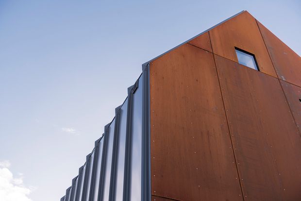 Close-up exterior architectural view of a sharply angled, contemporary building corner. Large rust-toned metal facade panels contrast with a rhythm of dark vertical glazed or metal fins along the side, with a small irregular window near the upper level. The image emphasizes the building envelope and geometric form against a clear sky.