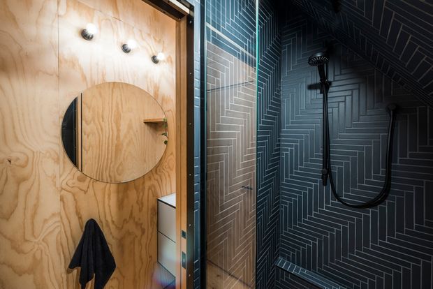 Compact contemporary bathroom showing a walk-in shower lined with dark grey herringbone tiles and black fixtures. Beside it is a warm plywood-clad vanity area with a round mirror, globe-style wall lights, timber shelving, and a small white drawer unit. The space has a strong contrast between warm timber and moody dark tile finishes.