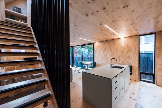 Open-plan timber-lined interior featuring a compact kitchen with a central island, integrated sink and dark cabinetry, adjacent dining area, and an open-riser staircase. Natural plywood walls and ceiling, timber flooring, black vertical stair screening, and generous glazing create a warm contemporary space.