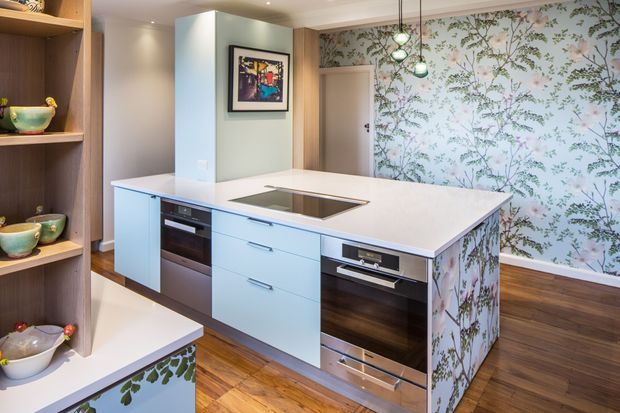 A bright contemporary kitchen centred on a large white island with pale blue cabinetry, integrated ovens, and a flush induction cooktop. The island and rear wall feature coordinated botanical-patterned wallpaper or printed panels, while timber shelving, hardwood flooring, and pendant lights add warmth and character.
