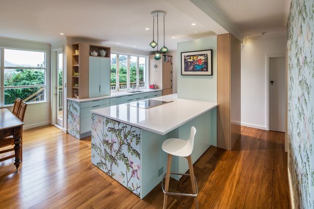 Bright, spacious kitchen with a large central island, integrated cabinetry, white benchtops, pale green joinery, patterned botanical wall panels, timber flooring, pendant lights, and abundant windows overlooking greenery. A dining area is partially visible at left.