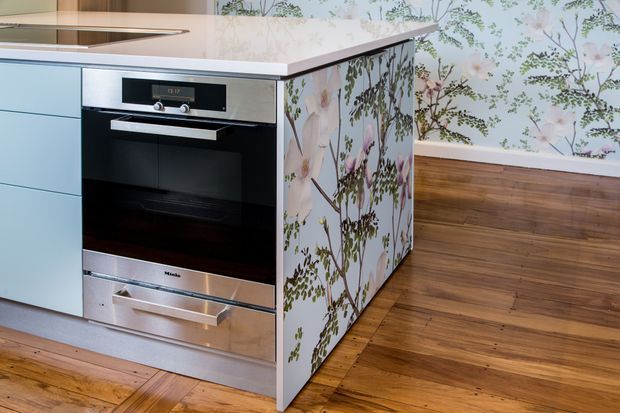 Close-up view of a contemporary kitchen island with an integrated black-and-stainless-steel oven, pale blue cabinetry, white benchtop, and a floral botanical patterned side panel. The kitchen sits over warm timber flooring, with matching botanical wallpaper visible on the wall behind.