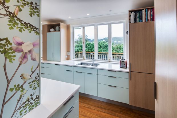Bright contemporary kitchen with pale blue-green handleless cabinetry, white benchtops, warm timber flooring, and a tall timber-toned storage unit with open book shelving. Large windows overlook a leafy landscape, while a botanical floral wall mural adds a decorative feature.