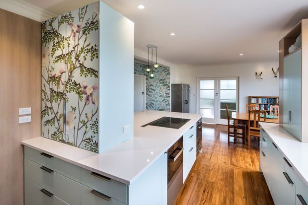 A bright open-plan kitchen with pale blue-grey built-in cabinetry, white stone benchtops, an integrated cooktop and oven, and a tall central island. A botanical floral feature wallpaper panel adds colour beside warm timber wall panelling. The kitchen opens toward a timber-floored dining area with a wooden table, chairs, shelving, and glazed exterior doors. Recessed ceiling lighting and pendant lights provide illumination.