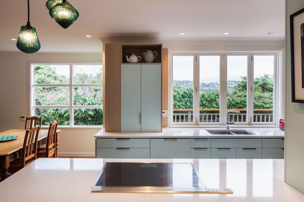 Bright contemporary kitchen with pale blue-grey cabinetry, white benchtops, an integrated induction cooktop, and a central timber pantry or storage unit. Large windows and glazed doors bring in abundant natural light and overlook greenery, while pendant lights hang above the adjacent dining area.