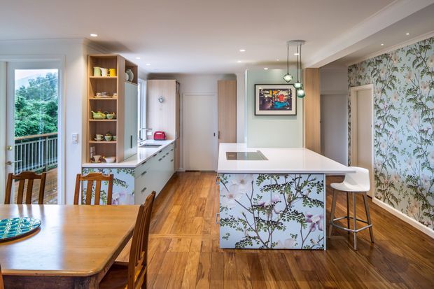 A bright open-plan kitchen and dining area with warm timber flooring and dining furniture. The kitchen features pale cabinetry, a white island with a floral botanical-patterned base, a matching floral wallpaper feature wall, open timber shelving, integrated appliances, and contemporary pendant lighting. A balcony or exterior greenery is visible through the glazed door.
