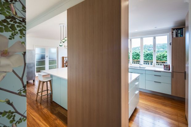 A bright contemporary kitchen viewed through a timber-lined opening, with warm timber flooring, pale cabinetry, a white island with bar stools, pendant lights, and large windows overlooking greenery. A floral feature surface is visible at the left edge, while tall timber cabinetry frames the kitchen.