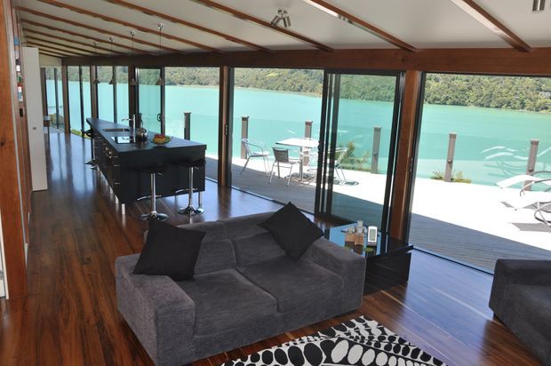 Open-plan waterfront living area with a dark grey sectional sofa in the foreground, a compact kitchen island with stools to the left, and full-height timber-framed glazing opening onto a timber deck. The room has dark polished timber flooring, exposed timber ceiling beams, and expansive views across turquoise water and wooded hills.